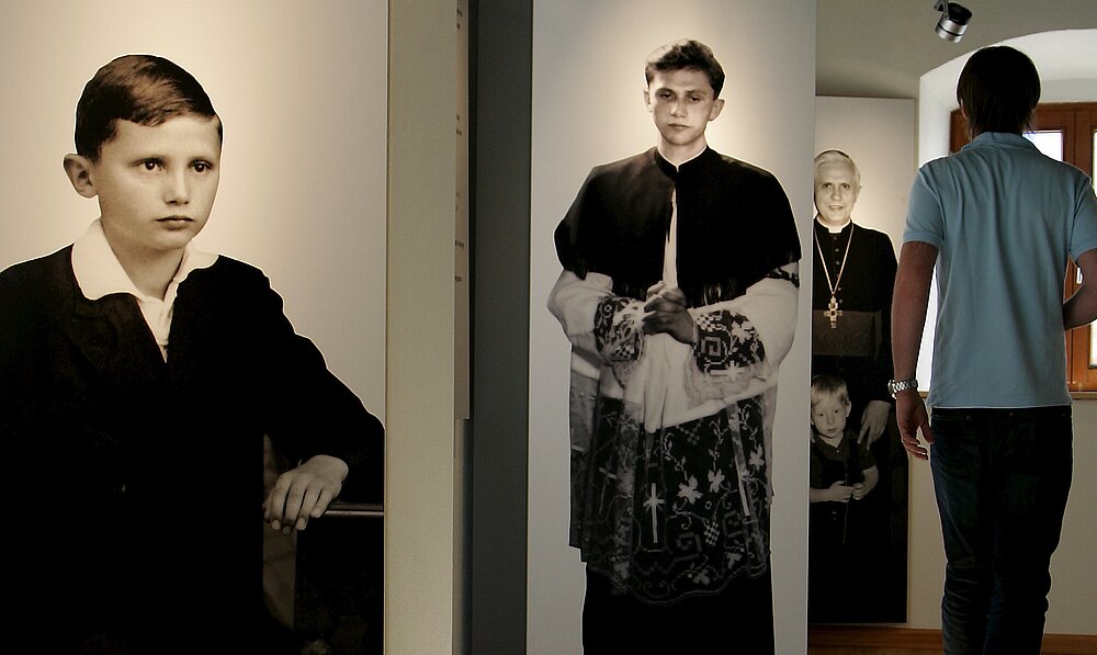 ARCHIV - Ein Mann läuft am Freitag (13.04.2007) in Marktl am Inn im Geburtshaus von Papst Benedikt XVI. an Fotos des Pontifex vorbei. Papst Benedikt XVI. hieß früher Joseph Ratzinger. Das Foto im Vordergrund zeigt den jungen Joseph Ratzinger. Es stammt aus einer Ausstellung. 