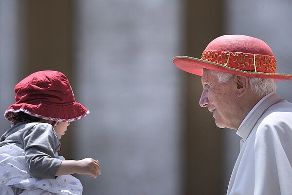 Papst Benedikt XVI. lächelt ein kleines Mädchen an. Beide tragen einen roten Hut.