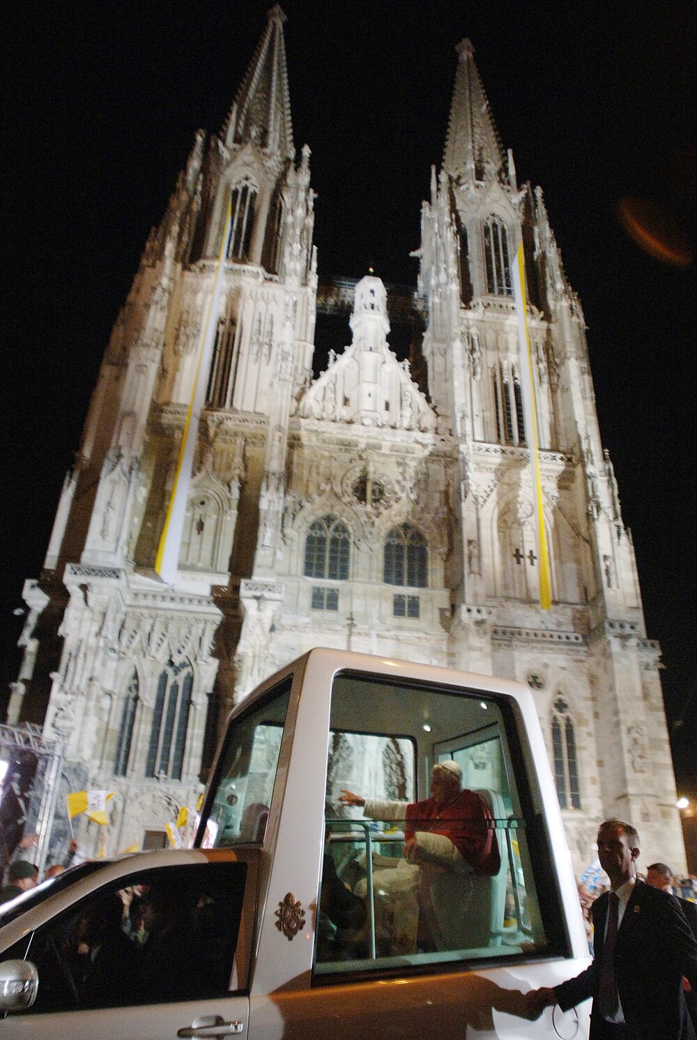 Papst Benedikt XVI sitzt im Papamobil und winkt. Im Hintergrund ist der Regensburger Dom zu sehen.