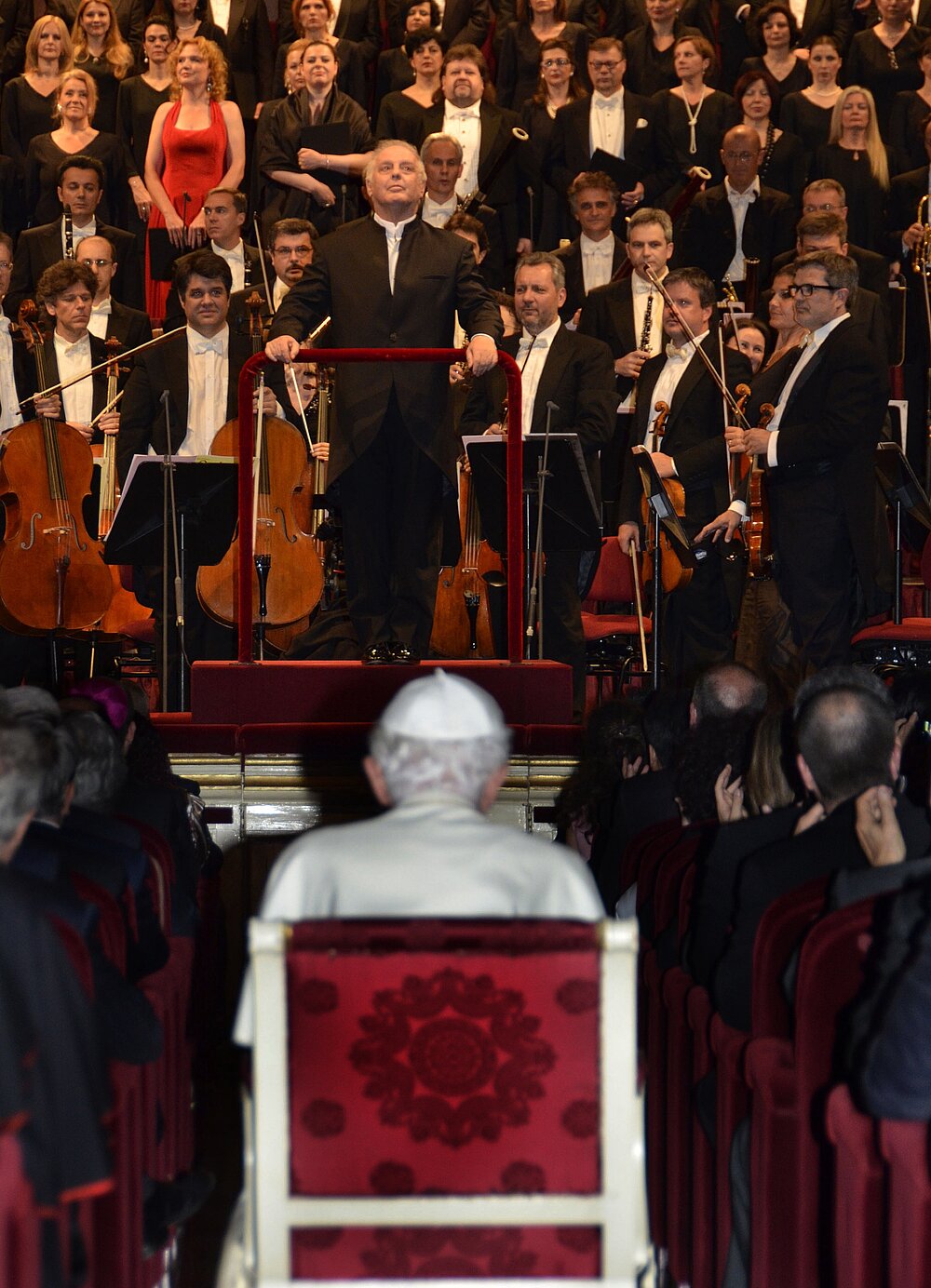 Papst Benedikt sieht man von hinten auf einem Stuhl sitzend und schaut auf eine Bühne mit Streichorchester.